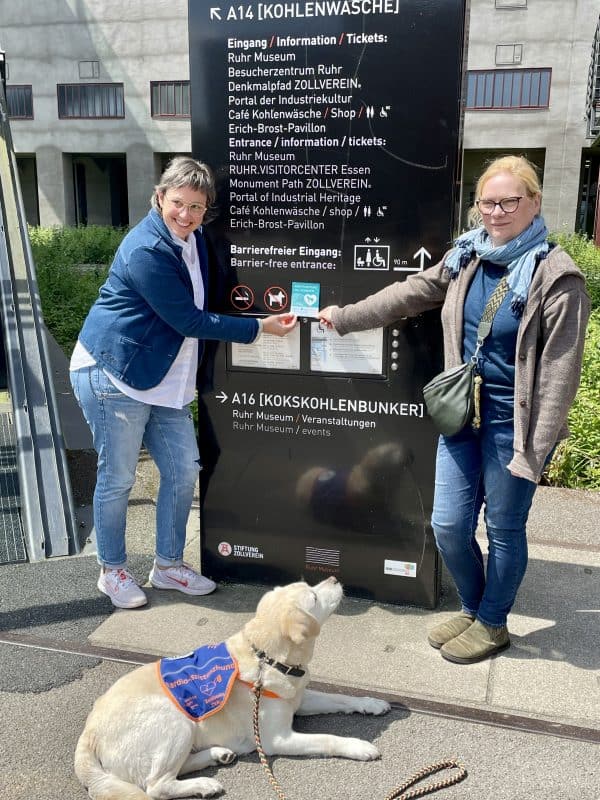 Frau Ladnar-Duckwitz steht mit der Botschafterin der Pfotenpiloten an der Infosäule des Besuchereinganges. Vor ihnen liegt der gelbe Labrador mit Kenndecke und schaut zu ihnen. Beide Menschen halten den Assistenzhund Willkommen Aufkleber in der Hand.