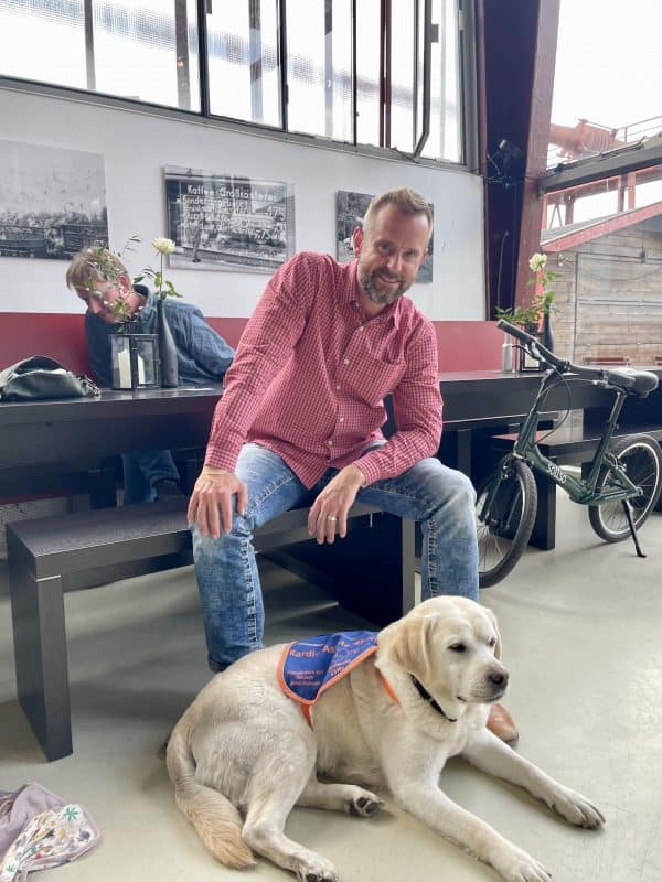 A man in jeans and a checked shirt is sitting on a bench.
It's the head of event management, Dirk Scheffler.
Mascha is lying in front of him.  