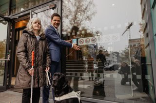 Regionspräsident Steffen Krach, Therese Fleßner mit ihrem Assistenzhund Folke kleben gemeinsam den Aufkleber „Assistenzhund Willkommen“ auf die Glastür.