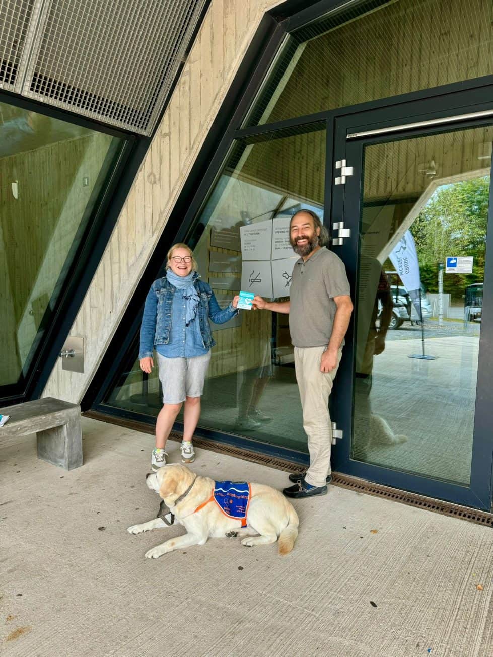 Herr Havlik steht mit einer Assistenzhundehalterin an der Eingangstür. Davor liegt der Assistenzhund. Die beiden Menschen halten den Aufkleber "Assistenzhund willkommen" gemeinsam fest.