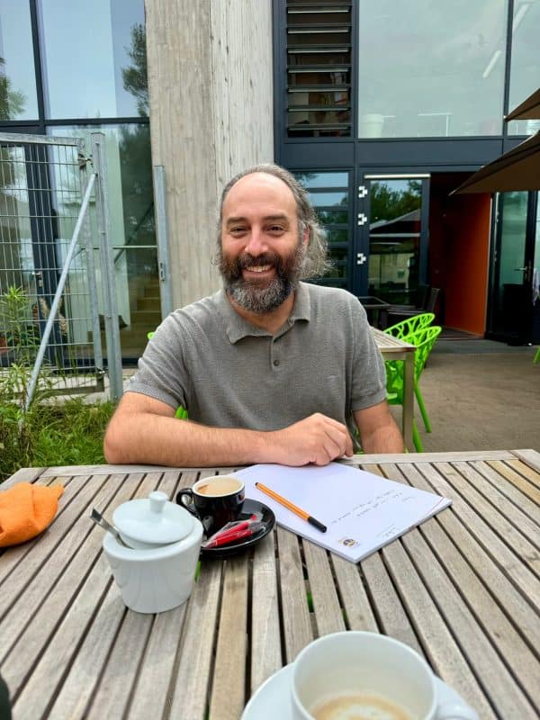 Ein Mann mittleren Alters sitzt am Tisch auf der Terrasse und strahlt in die Kamera.