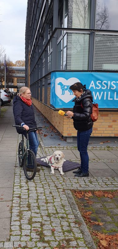 Eine Assistenzhundhalterin sitzt auf dem Laufrad, ihr Assistenzhund liegt neben ihr auf einer Decke, eine Reporterin interviewt die Assistenzhundhalterin.