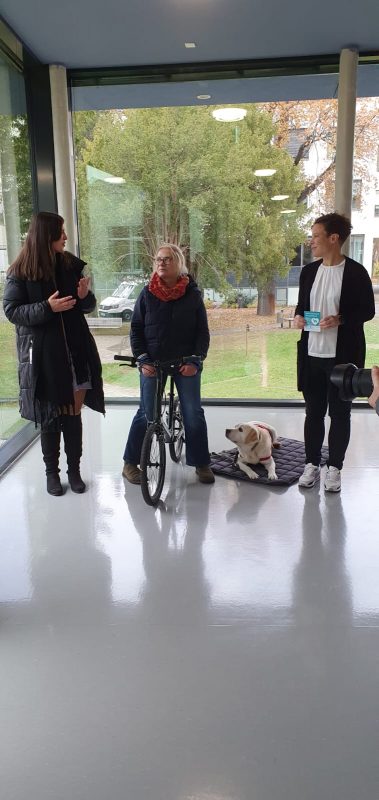 Frau Dr. Denninger und Frau Herrschuh stehen mit dem Assistenzhundteam vor einer Glasfront bei einer gemeinsamen Pressekonferenz