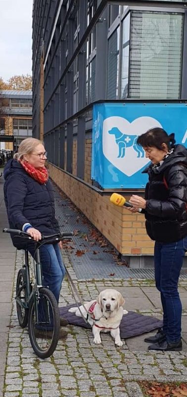 Eine Assistenzhundhalterin sitzt auf dem Laufrad, ihr Assistenzhund liegt neben ihr auf einer Decke, eine Reporterin interviewt die Assistenzhundhalterin.